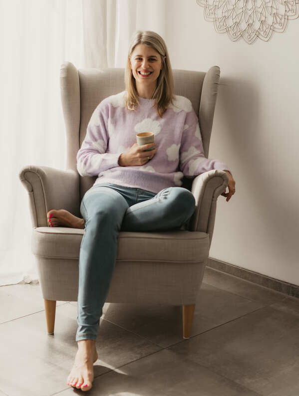 Finja Wolkenstein sitzt mit gekreuzten Beinen auf ihrem Sessel und hält einen Kaffeebecher in der Hand.